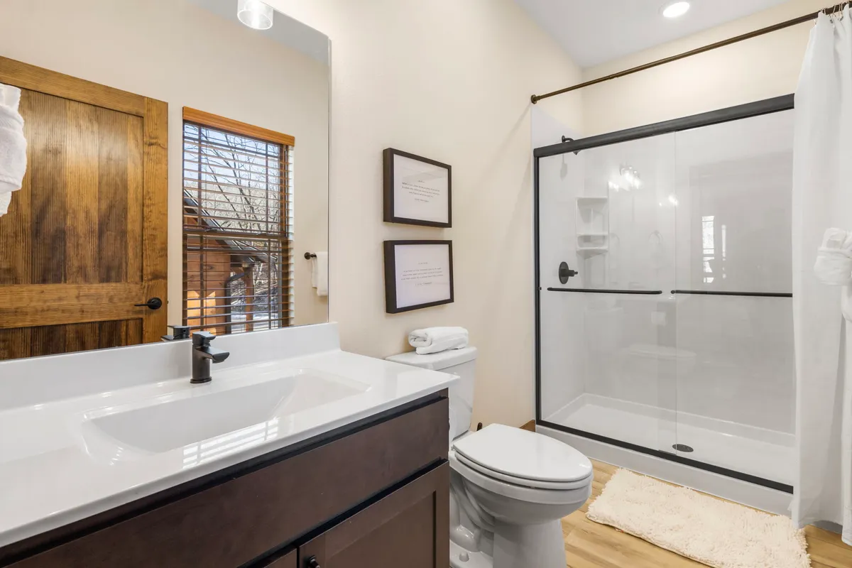 A modern ensuite bathroom with a glass shower, a wide sink, a large mirror, and natural light filtering in. Subtle wooden accents and framed prints add warmth and modern flair to the space.