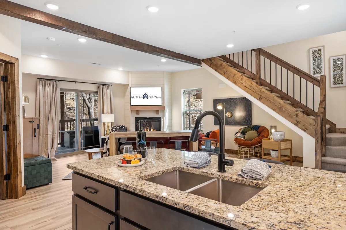 A homey interior with a granite kitchen island, comfortable seating area by the fireplace, and a staircase leading to other areas. The space is designed for both relaxation and group activities, with a clear view of the interconnected living, dining, and kitchen spaces.