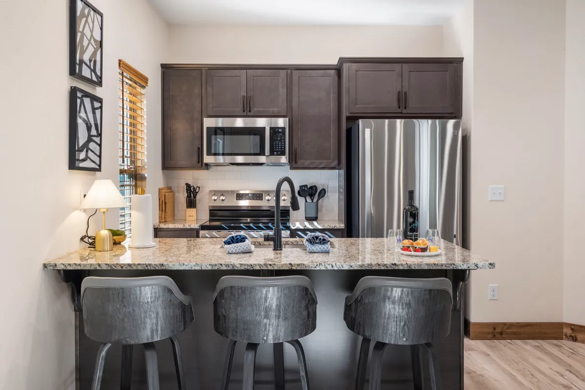 A contemporary kitchen scene with a granite countertop, modern bar stools, stainless steel appliances, and tasteful artwork on the wall. The space is clearly designed for both practical cooking and socializing, with room for guests to gather and enjoy meals together.