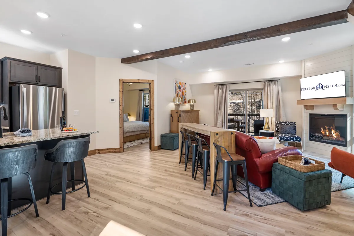A warm, open-concept living space featuring a kitchen island, modern fireplace, dining area, and an inviting living area with plush seating. Large windows fill the room with natural light and offer views of the surrounding woods, creating a seamless indoor-outdoor connection.