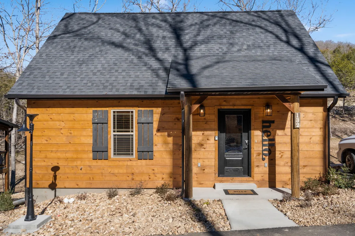 The inviting wooden exterior of a modern cabin with black shutters, a welcoming front door, and a cozy shaded porch. Lush greenery surrounds the property, hinting at nearby nature trails and a peaceful, secluded setting.