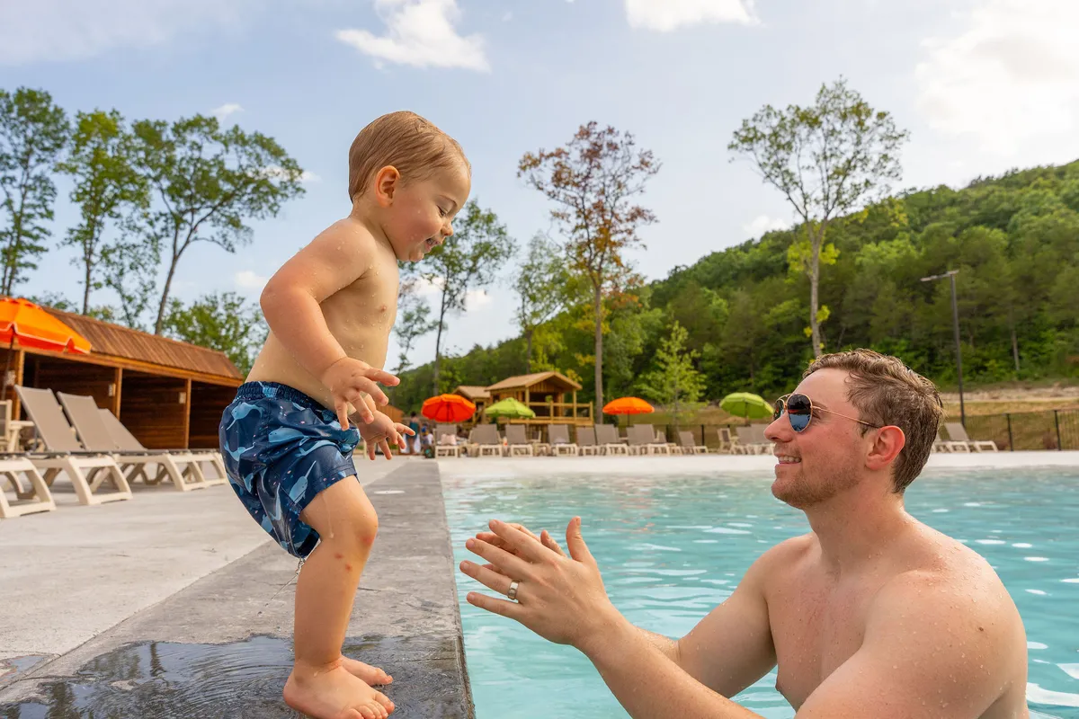 A child leaps joyfully into an outdoor swimming pool while a father waits with open arms, surrounded by colorful umbrellas and the backdrop of the resort. The scene exudes carefree fun, laughter, and family togetherness.