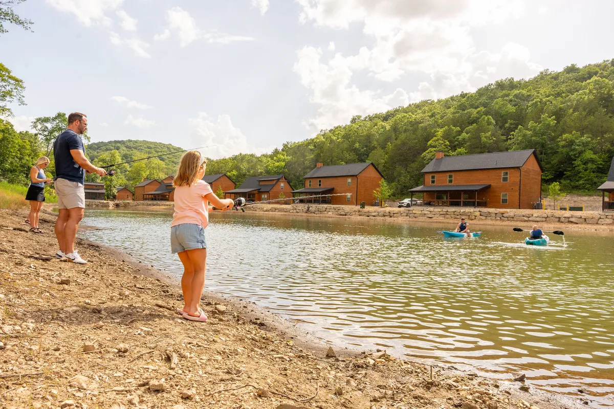 This exterior scene captures a family fishing and kayaking by a serene lake, with cozy cabins visible in the background. The lush greenery and calm water set a playful and adventurous tone, emphasizing family bonding and outdoor activities.