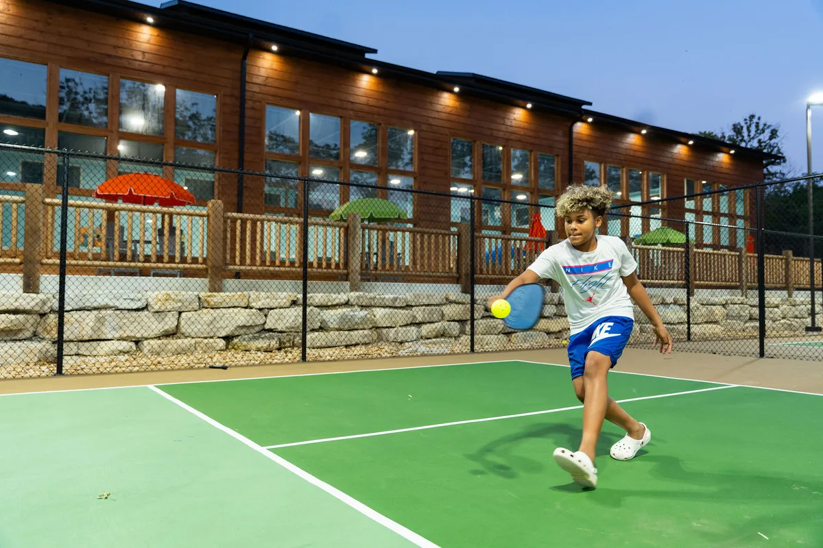 This lively exterior scene captures people playing pickleball on a vibrant court beside a wooden building, with colorful umbrellas and a pool in the background, all set amid green trees. The action and joyful mood convey friendly competition and group bonding.